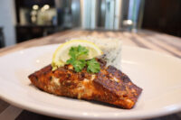 Blackened Salmon fillet with a lemon wedge and parsley garnish beside herbed rice on a white plate.