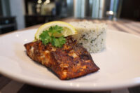 Blackened Salmon fillet with lemon wedge and parsley, served beside herbed rice on a white plate.