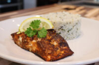 Blackened Salmon fillet with a dark spice crust, topped with parsley and lemon, beside herbed rice on a white plate.