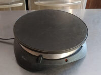 Crepes An electric crepe maker with a round black cooking plate sits unused on a stainless steel counter, the appliance used to prepare Crepes.