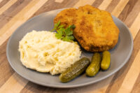 Dutch Schnitzel (Holandský Řízek) Golden fried Dutch Schnitzel (Holandský Řízek) with mashed potatoes, parsley, and pickles on a gray plate.