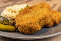 Dutch Schnitzel (Holandský Řízek) Two golden fried Dutch Schnitzel (Holandský Řízek) cutlets on a grey plate with gherkins and a shredded white side.