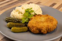 Dutch Schnitzel (Holandský Řízek) Golden fried Dutch Schnitzel (Holandský Řízek) served with mashed potatoes, pickles, and parsley on a gray plate.