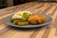 Dutch Schnitzel (Holandský Řízek) Golden breaded Dutch Schnitzel (Holandský Řízek) on a grey plate with mashed potatoes and pickles.