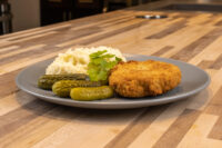 Dutch Schnitzel (Holandský Řízek) Golden breaded Dutch Schnitzel (Holandský Řízek) on a grey plate with mashed potato and pickles.