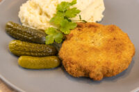 Dutch Schnitzel (Holandský Řízek) Golden breaded Dutch Schnitzel (Holandský Řízek) on a grey plate with mashed potato, gherkins and parsley.
