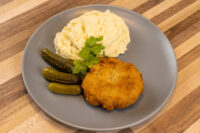 Dutch Schnitzel (Holandský Řízek) Golden breaded Dutch Schnitzel (Holandský Řízek) on a grey plate with mashed potatoes and gherkins.