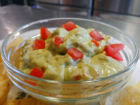 Guacamole Glass bowl of Guacamole topped with creamy sauce and diced tomato, served with tortilla chips.