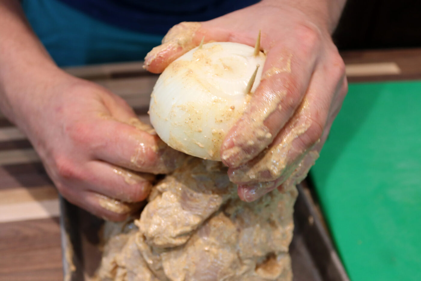 Hands press an onion pierced with toothpicks into a mound of yogurt-marinated Chicken Gyros meat.