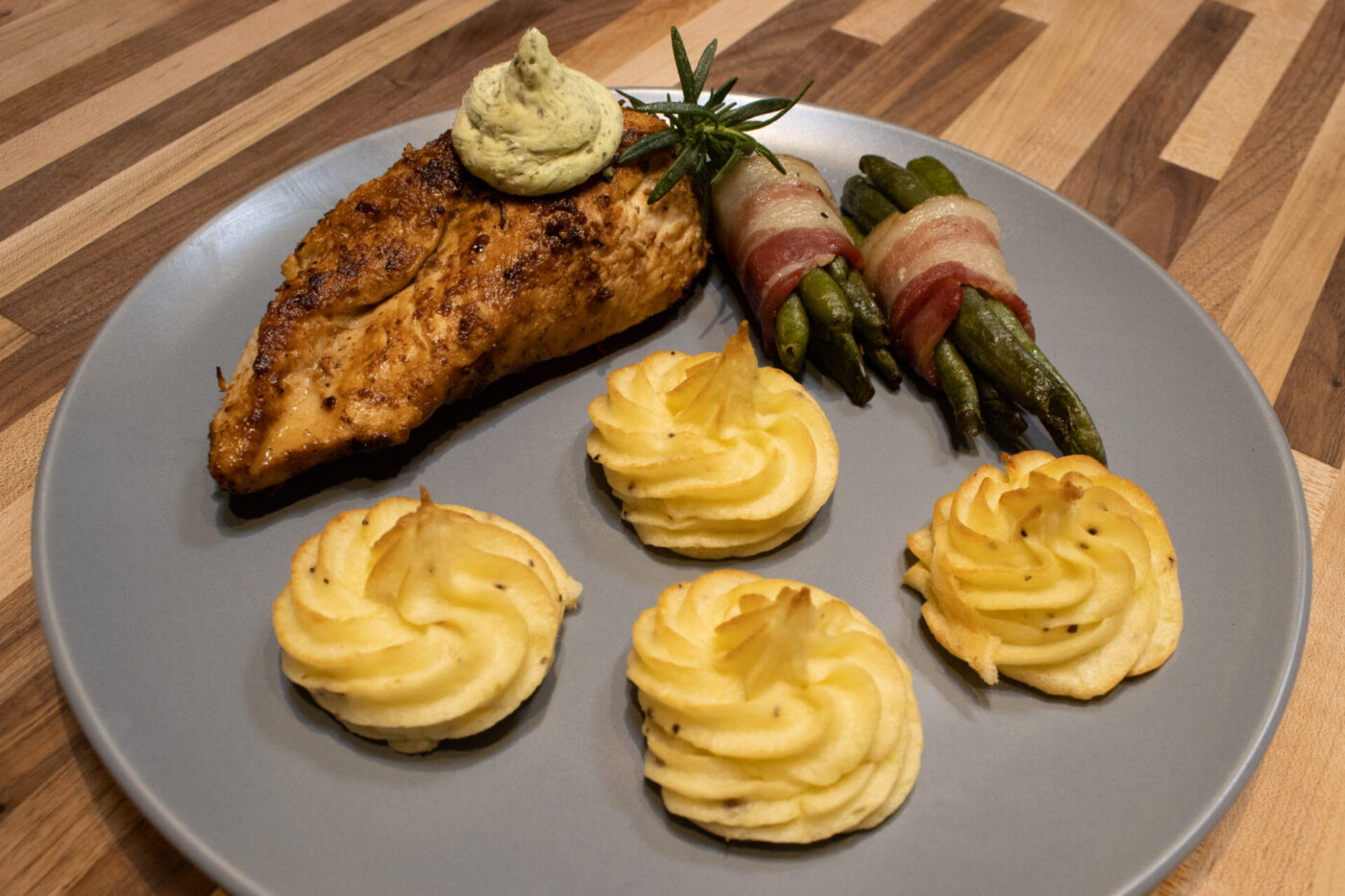 Rosemary Chicken Breasts Plated Rosemary Chicken Breasts with herb butter, duchess potatoes and bacon-wrapped green beans on a grey plate.