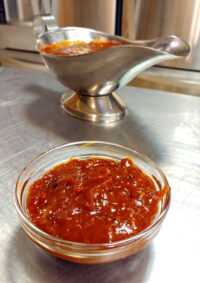 BBQ Sauce Glass bowl of chunky BBQ Sauce with a metal gravy boat of the same sauce behind it on a steel counter.