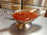 BBQ Sauce Silver gravy boat filled with thick reddish-orange BBQ sauce on a stainless steel counter.