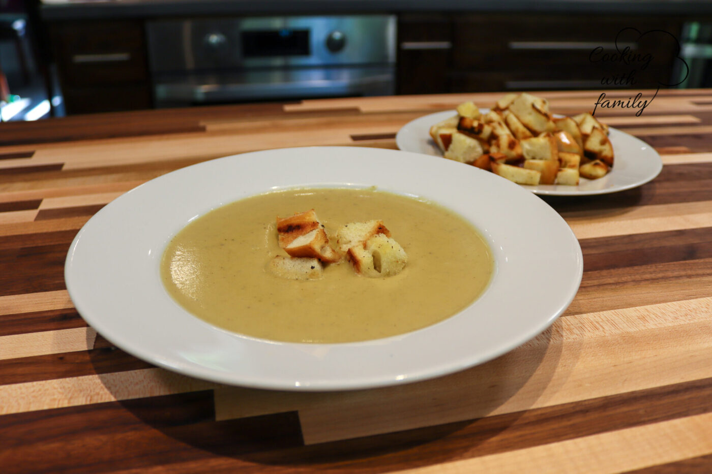 Broccoli Soup Bowl of creamy Broccoli Soup topped with croutons, extra croutons on a plate behind it on a wooden table.