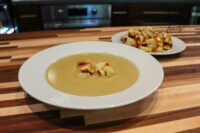 Broccoli Soup Bowl of creamy Broccoli Soup topped with croutons, extra croutons on a plate behind it on a wooden table.