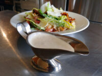 Caesar Dressing (easy version) A metal gravy boat of Caesar Dressing (easy version) sits in front of a Caesar salad on a steel table.