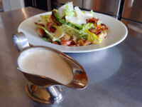 Caesar Dressing (easy version) Creamy Caesar Dressing filling a metal gravy boat, set in front of a bacon and romaine salad.