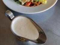Caesar Dressing (easy version) Creamy Caesar Dressing with flecks of seasoning pooled in a small metal creamer, salad plate blurred behind.