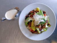Caesar Salad Caesar Salad with romaine, shaved parmesan, bacon, and tomato on a white plate, dressing in a metal pitcher nearby.