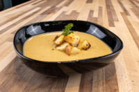 Black bowl of creamy Cauliflower Soup topped with croutons and parsley on a wood table.