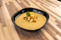 Black square bowl of Cauliflower Soup topped with croutons and parsley, on a wooden table.