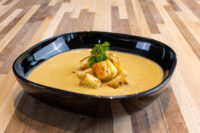 Bowl of creamy Cauliflower Soup topped with roasted potato cubes and a parsley sprig on a wood table.