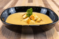 Black oval bowl of Cauliflower Soup topped with toasted croutons and a parsley sprig on a wooden table