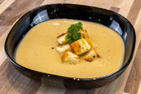 Black bowl of Cauliflower Soup topped with toasted croutons and a parsley sprig on a wooden table.