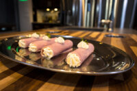 Creamy Horseradish Ham Rolls Five Creamy Ham Rolls piped with white filling and garnished with herbs, arranged on an oval metal tray on a striped wooden table