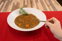 White bowl of Czech Lentil Soup Čočková Polévka with lentils and carrot slices, garnished with rosemary, on a red cloth.