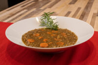 White bowl of Czech Lentil Soup Čočková Polévka with carrot slices, topped with a fresh herb sprig, on red cloth.