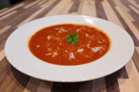 Czech Tomato Soup with Rice White bowl of Czech Tomato Soup with Rice, garnished with parsley, on a striped wooden table.