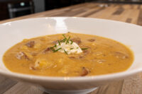 A bowl of Frankfurt Soup Frankfurtská Polévka with diced potato, sliced kielbasa, a dollop of sour cream, and chives, set on a wooden table.