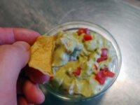 Guacamole Hand dips a tortilla chip into a bowl of guacamole with diced tomato on a steel counter.