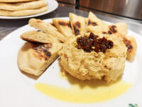 Hummus Round mound of hummus topped with dark relish and olive oil, ringed by grilled pita wedges on a white plate.