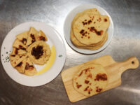 Hummus Bowl of hummus topped with a dark red garnish and olive oil, ringed by pita wedges, with extra flatbreads on the side.