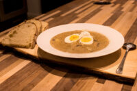 White bowl of creamy Kulajda Soup topped with two soft-boiled egg halves, served with bread on a wooden board.