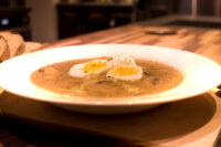 White bowl of creamy Kulajda Soup topped with two egg halves and black pepper, bread beside it.