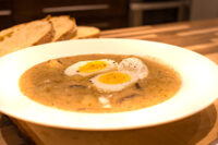 White bowl of Kulajda Soup with mushroom pieces, dill, and soft-boiled egg halves, sliced bread behind.