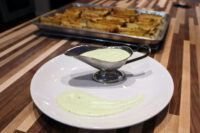 Pale green aioli swirled on a white plate and pooled in a metal gravy boat, potato wedges on a tray behind.