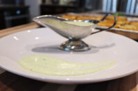 A swipe of pale green Mediterranean Aioli on a white plate beside a metal gravy boat filled with the same sauce.