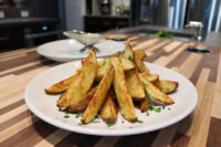 Oven-Roasted Potato Wedges Golden Oven-Roasted Potato Wedges piled on a white plate, topped with chopped green chives, on a wooden counter.