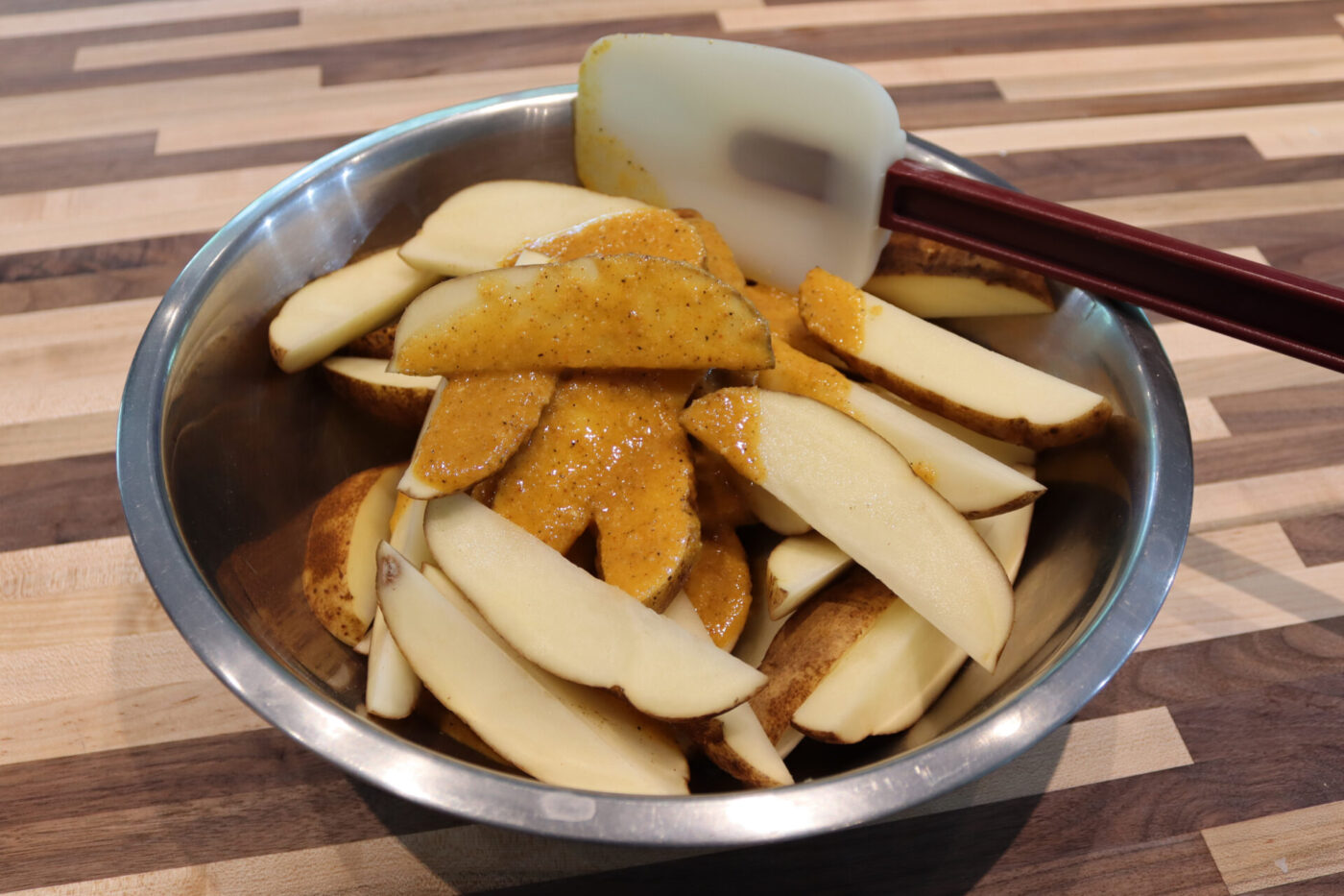 Potato wedges for Oven-Roasted Potato Wedges coated in orange spice paste in a metal bowl with a spatula.