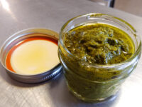 Pesto A glass jar filled with green pesto, its metal lid resting on a steel counter beside it.