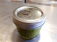 Pesto Small glass jar labeled 'Pesto 2oz' filled with green pesto, sitting on a steel countertop.