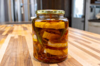 Jar of Pickled Brie Cheese Nakladany Hermelin with onion strips and herbs steeped in oil on a wooden counter.