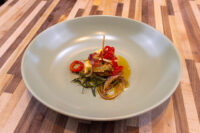 Plated Nakladaný Hermelín with chilli slices, herb sprigs and crispy onion in orange oil, pale bowl on wood table.