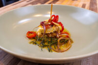A piece of Pickled Brie Cheese Nakladaný Hermelín topped with red pepper and onion rings, resting on herbs in oil.