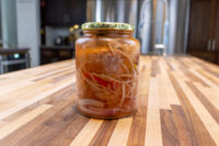 Pickled Sausage Utopenec Glass jar of Pickled Sausage Utopenec with onion strands and red pepper in brine on a wood counter.