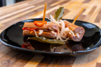 Pickled Sausage Utopenec Two skewered rolled sausages of Pickled Sausage Utopenec with pickled onions, red pepper, and bay leaf on a black plate.