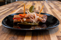 Pickled Sausage Utopenec Two browned sausages of Pickled Sausage Utopenec topped with onion, pepper, and bay leaf on a black plate.